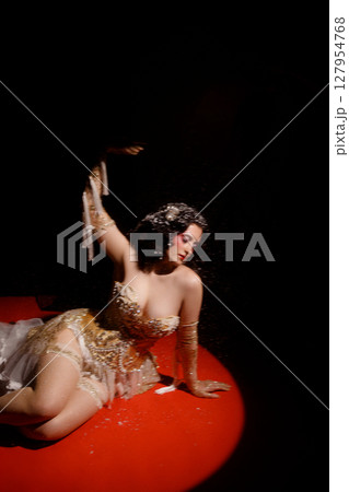 Woman in vintage corset lying on red stage floor under hanging beads on dark background 127954768