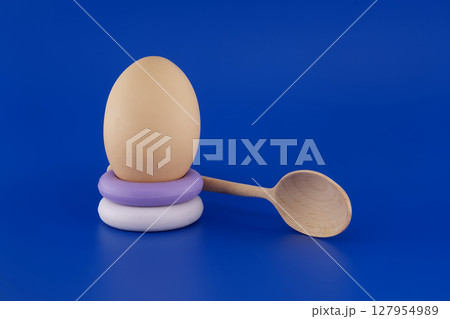 Egg balanced on stacked soap rings alongside a wooden spoon on a blue backdrop. Egg balanced on stacked soap rings alongside a wooden spoon on a blue backdrop. 127954989