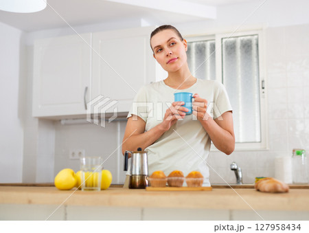 Dreamy young woman standing in home kitchen with cup of coffee Dreamy young woman standing in home kitchen with cup of coffee 127958434