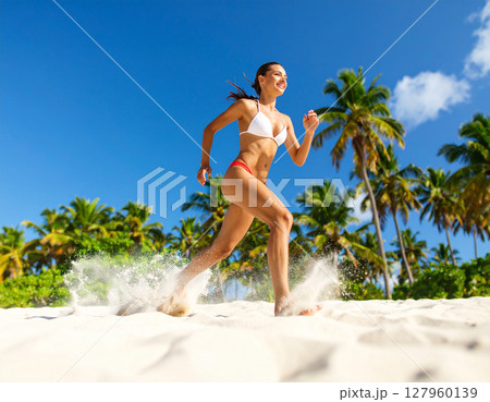 AI generated dynamic low angle shot of woman in red and white bikini running across the camera with sand kicking up, palm trees in background under bright blue sky creating energetic summer atmosphere 127960139