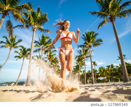 AI generated dynamic low angle shot of woman in red and white bikini running across the camera with sand kicking up, palm trees in background under bright blue sky creating energetic summer atmosphere 127960140