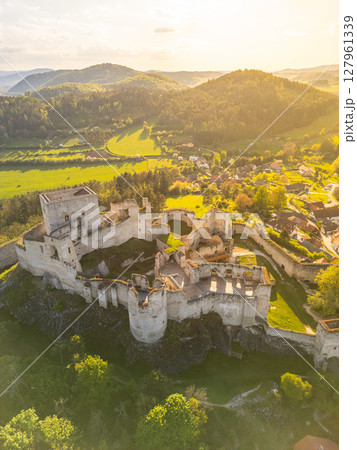 Aerial view reveals the majestic ruins of Rabi Castle surrounded by lush green hills and fields in Czechia. The landscape showcases the rich history and beauty of the region during sunset. 127961339