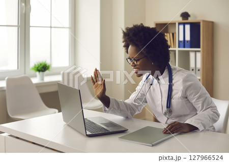 Young dark-skinned female doctor provides an online consultation to the patient via video link. 127966524