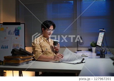Focused man in glasses holding coffee while working overtime in a modern office 127967201