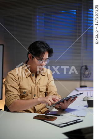Focused office worker analyzing data on digital tablet during overtime hours in an office 127967208
