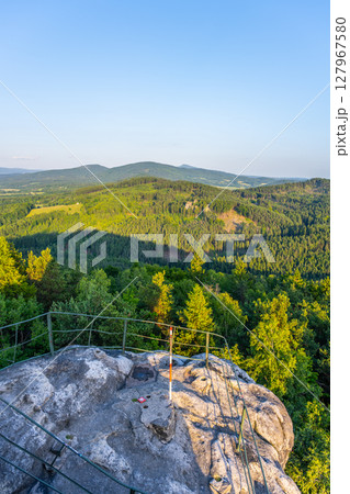 Jested Ridge is bathed in golden light as the sun sets behind the Lusatian Mountains. The sandstone cliffs rise above lush green forests, creating a breathtaking evening view. 127967580