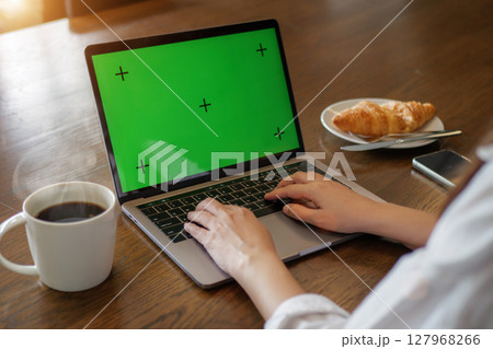 laptop blank green screen mockup. woman using laptop computer, hand typing on keyboard at home for work with hot coffee, croissant bakery and mobile phone on the wooden table in the morning. 127968266