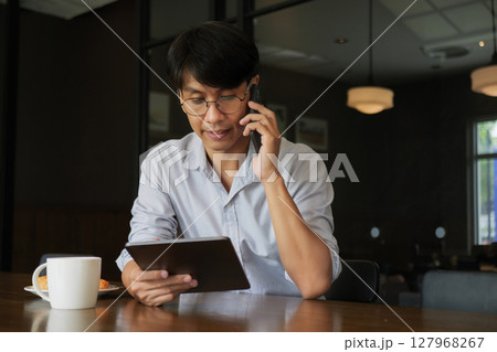 Asian man wearing glasses talking on the phone while looking at information on a tablet in his hand for work in the morning with hot coffee and croissant bakery on the wooden table at working space. 127968267