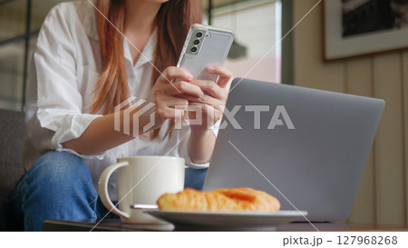 front view of woman using phone and laptop computer at coffee shop for work in the morning with hot coffee, croissant bakery and mobile phone on the table. anonymous face 127968268