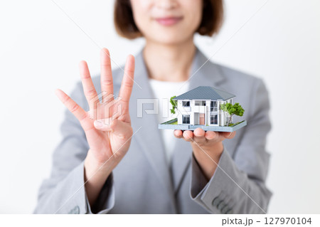 家の住宅模型を持って4本指を立てるグレーのスーツ姿のミドル女性 127970104