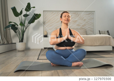 Yoga mindfulness meditation. Woman practicing yoga in living room at home. Plus size female sitting in lotus pose meditating smiling relaxing indoor. Girl doing breathing practice. Yoga at home 127970300