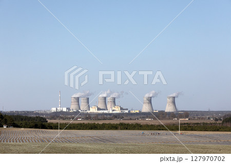 view of a nuclear power plant. View of smoking chimneys of nuclear power plant, power lines and forest, under blue sky with white clouds 127970702