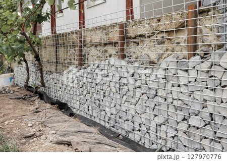 Stone fence in steel mesh and green lawn. Close up of gabion with tightly packed stones of varying sizes and textures. Combination of natural materials and industrial design. Stone fence in steel mesh and green lawn. Close up of gabion with tightly packed stones of varying sizes and textures. Combination of natural materials and industrial design. 127970776