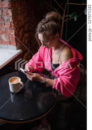 A stylish woman enjoying her coffee while scrolling on her phone in a warm cafe setting. A stylish woman enjoying her coffee while scrolling on her phone in a warm cafe setting. 127973182