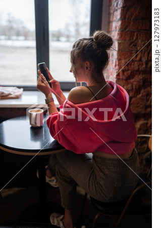 Charming woman with beautiful smile reading good news on mobile phone during rest in coffee shop, happy Caucasian female watching her photo on cell telephone while relaxing in cafe during free time  127973183