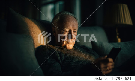 Elderly man sitting on sofa, illuminated by smartphone screen, in dimly lit room 127973621