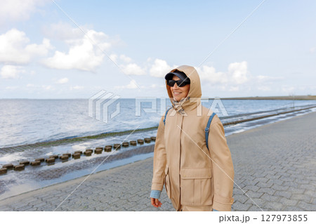 Smiling happy woman walking by Nord sea coast wear beige coat sunglasses enjoys windy day cloudy sky background. Female person have fun hiking adventure travel at Baltic sea seaside Smiling happy woman walking by Nord sea coast wear beige coat sunglasses enjoys windy day cloudy sky background. Female person have fun hiking adventure travel at Baltic sea seaside 127973855