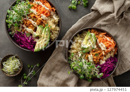 Two colorful bowls filled with quinoa, shredded carrots, sliced avocado, purple cabbage, sprouts, and creamy dressing. A rustic cloth and scattered greens enhance the earthy, fresh presentation. Two colorful bowls filled with quinoa, shredded carrots, sliced avocado, purple cabbage, sprouts, and creamy dressing. A rustic cloth and scattered greens enhance the earthy, fresh presentation. 127974451