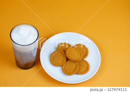 A refreshing glass of iced coffee with a foamy top and a plate of golden-brown biscuits, all against a vibrant orange backdrop. A refreshing glass of iced coffee with a foamy top and a plate of golden-brown biscuits, all against a vibrant orange backdrop. 127974513