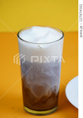 A close-up of a tall glass of iced coffee with a creamy white foam top, showing distinct layers of brown and white liquid against a bright orange background. 127974582