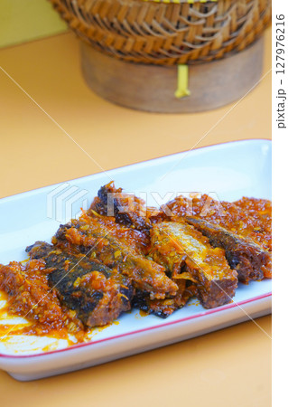A rectangular white plate holds several pieces of fried mackarel tuna fish in sambal balado or a rich, spicy chilli sauce, set against a plain yellow background. 127976216