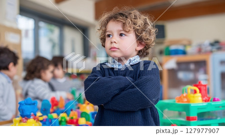 Pensive young boy with curly hair in a classroom with other students, looking thoughtful. He is standing near colorful toys with arms crossed. 127977907