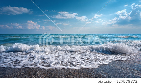 Waves crashing on the shore with foam. Ocean view with bright blue water under a sky with fluffy white clouds, creating a peaceful coastal scene. 127978872