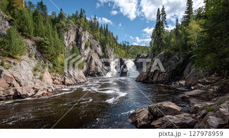 Scenic waterfall cascading into river below. Rocks and evergreen trees surround the falls on a sunny day with blue skies. Serene nature shot. 127978959