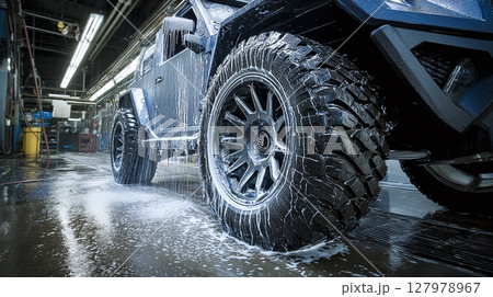 Vehicle is getting a cleaning with soap and water. The soap runs off the tires and frame. The car is black and has big wheels and tires. 127978967