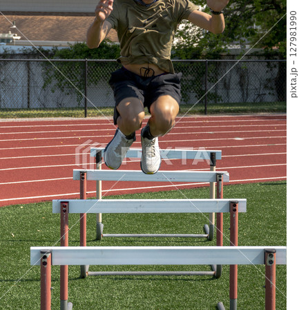 Athlete Practices Jumping Over Hurdles next to a Track on a Turf Field 127981990