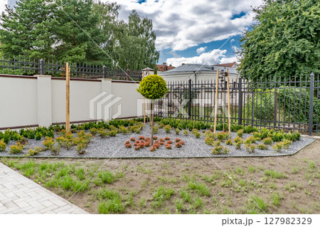 Back and front yard cottage garden, flowering plant and green grass lawn, brown pavement and orange brick wall, evergreen trees on background, in good care maintenance landscaping in park 127982329