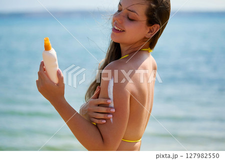 Beautiful woman enjoying a sunny day at the beach while applying sunscreen for skin protection and healthy tanning. Skincare concept 127982550