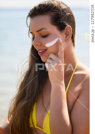 Beautiful woman enjoying a sunny day at the beach while applying sunscreen to protect her skin from harmful UV rays 127982556