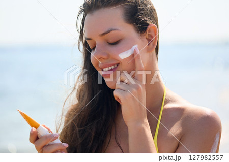 Beautiful woman enjoying a sunny day at the beach while applying sunscreen to protect her skin from harmful UV rays. Skincare concept 127982557