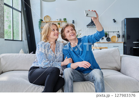 Middle aged mother and teenage son sitting together at home taking selfie photo on smartphone 127983376