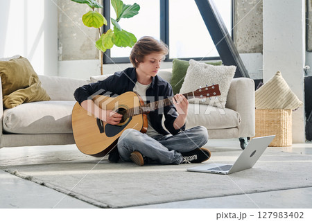 Teenage boy with acoustic guitar and laptop, having online music lesson 127983402