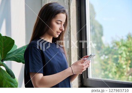 Teenage girl using smartphone standing at home near window, copy space Teenage girl using smartphone standing at home near window, copy space 127983710