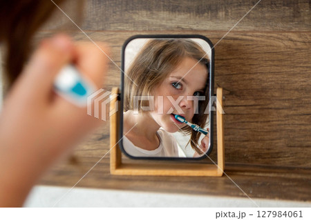 Oral hygiene, healthy teeth and care. Girl brushing teeth with toothbrush and looking in mirror in bathroom interior in the morning, closeup, empty space. High quality photo Oral hygiene, healthy teeth and care. Girl brushing teeth with toothbrush and looking in mirror in bathroom interior in the morning, closeup, empty space. High quality photo 127984061