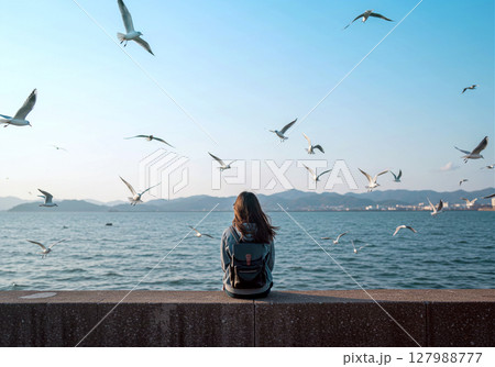 堤防に座り海鳥を見つめる少女の後ろ姿 127988777
