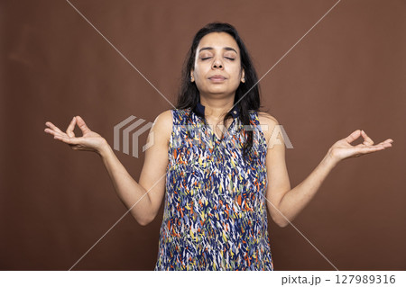 Beautiful woman with closed eyes and arms outstretched, standing in meditation against brown background. Portrait of Indian female individual immersed in a peaceful meditation session. 127989316