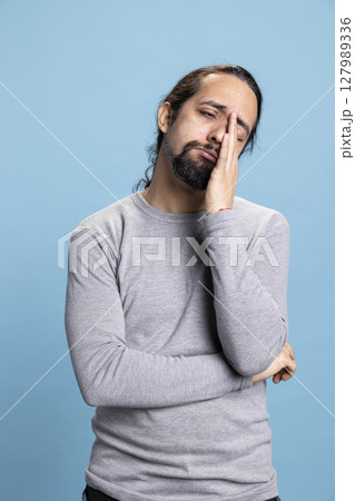 Sleepy male person dealing with burnout and fatigue while he stands in the studio, having a diversity of emotions. Young guy being tired and almost falling asleep against blue background. 127989336