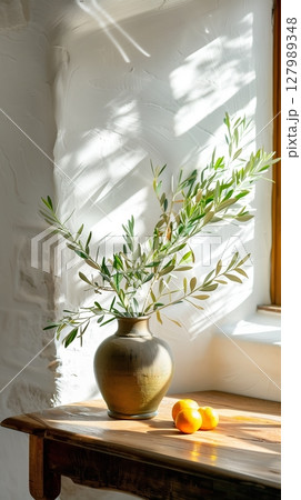 Rustic earthenware vase holding olive branches sitting alongside oranges near sunlit window, evoking warm Mediterranean ambiance on weathered wooden surface,Mediterranean ambiance 127989348