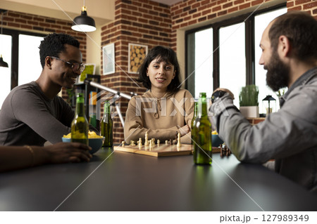 Caucasian woman with arms crossed, sitting in the middle of male friends during chess match in brick wall room. Selective focus on young lady looking at camera as others enjoy board game session. Caucasian woman with arms crossed, sitting in the middle of male friends during chess match in brick wall room. Selective focus on young lady looking at camera as others enjoy board game session. 127989349