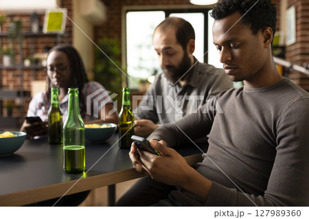 Close up of african american man using smartphone, with bottle of beer nearby on table. Black male individual engages in online browsing while sitting with friends in brick walled room. 127989360