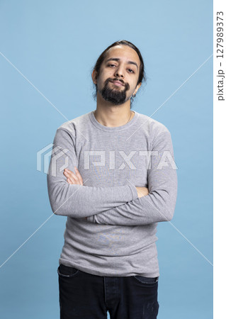 Young adult in casual wear standing with his arms crossed on camera, posing with confidence against a blue background. Man with a beard and long hair, calm natural attitude. Young adult in casual wear standing with his arms crossed on camera, posing with confidence against a blue background. Man with a beard and long hair, calm natural attitude. 127989373