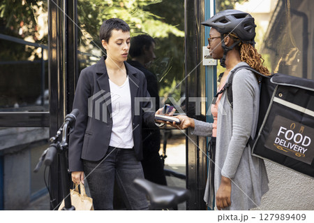 Black female worker accepts electronic payment from customer with a card on pos terminal. Courier ensuring transaction finalizes the purchase of fast food order from restaurant delivery. Black female worker accepts electronic payment from customer with a card on pos terminal. Courier ensuring transaction finalizes the purchase of fast food order from restaurant delivery. 127989409