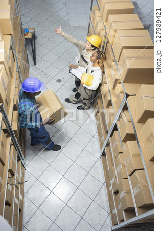 Multiethnic group of workers perform operational tasks in a depot, clean and structured factory space. Stacked boxes to handle the logistics chain, manual work unit in warehouse. Multiethnic group of workers perform operational tasks in a depot, clean and structured factory space. Stacked boxes to handle the logistics chain, manual work unit in warehouse. 127989421