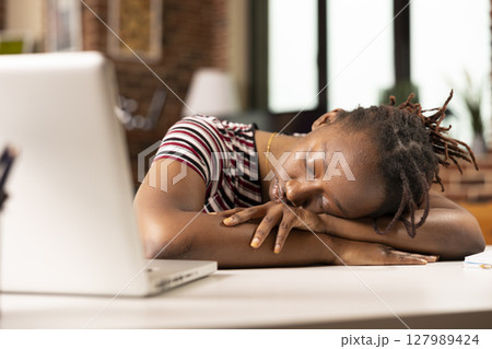 Black female entrepreneur lies on desk in home office, taking quick nap after demanding morning of work. Tired remote worker rests near laptop, overwhelmed by exhaustion from completing tight 127989424