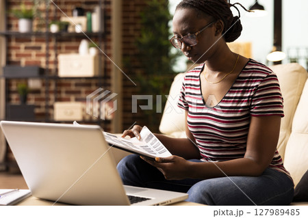 Determined female entrepreneur carefully reviewing printed documents during remote work session. Focused black woman flips through clipboard, preparing notes for upcoming project presentation. Determined female entrepreneur carefully reviewing printed documents during remote work session. Focused black woman flips through clipboard, preparing notes for upcoming project presentation. 127989485