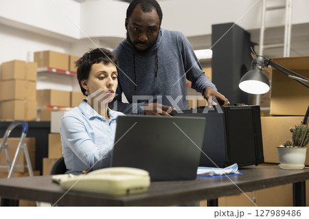 Diverse employees at small business fulfillment center manage local shipping and inventory processes, working together in a warehouse full of taped boxes and storage shelving units. 127989486